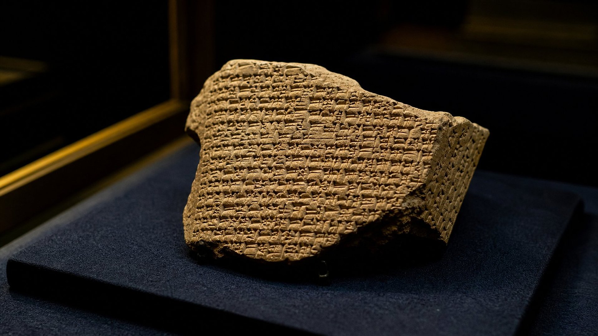 Gros plan d’un fragment de tablette d’argile brisé, couvert de signes cunéiformes serrés, posé sur un socle de velours bleu nuit dans une vitrine.