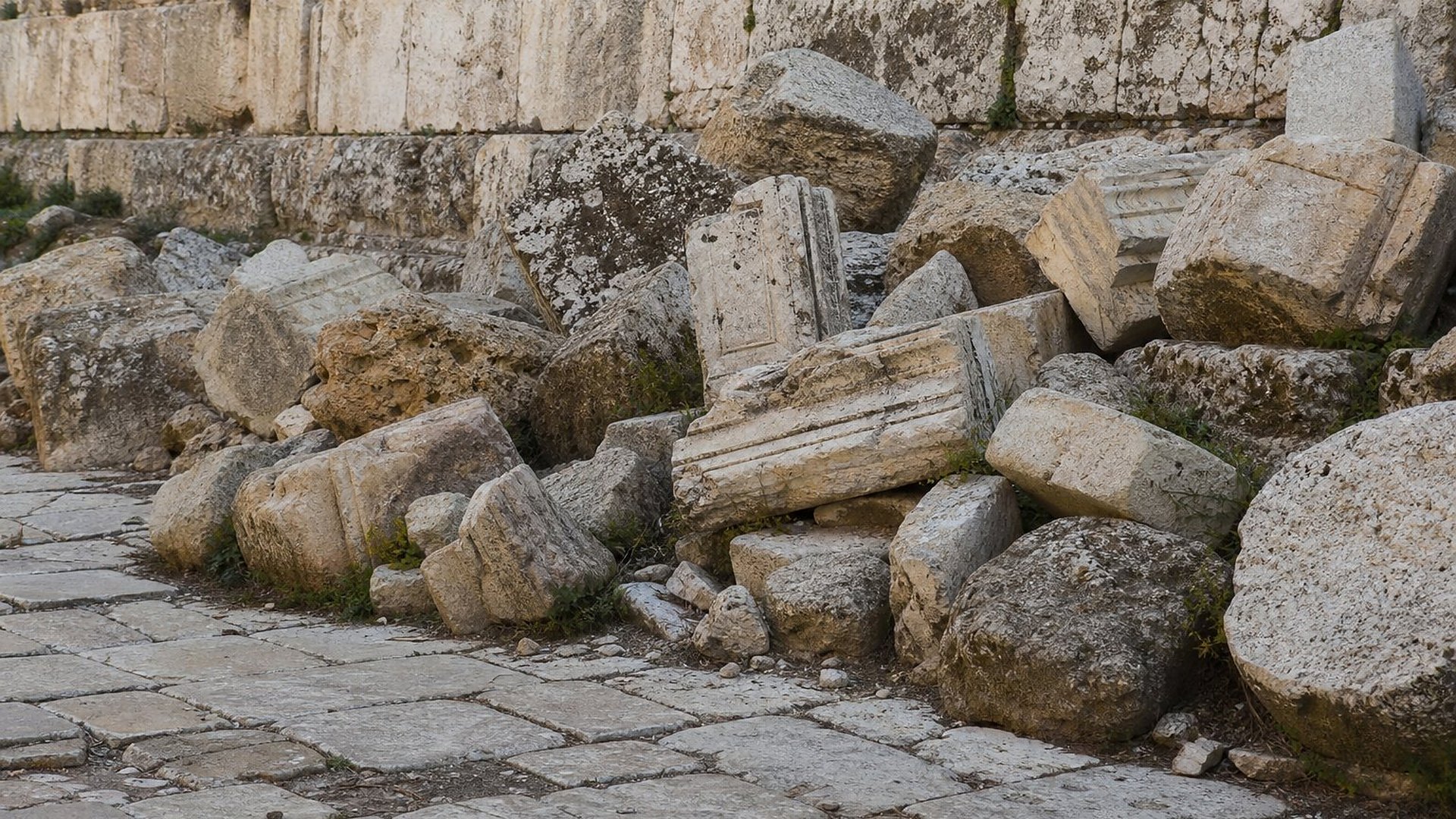 Au pied d’un mur fait d’énormes blocs de calcaire taillés, un amas de grosses pierres et de fragments sculptés, brisés et renversés, recouvre une rue antique pavée de grandes dalles ; de petites plantes poussent entre les blocs.