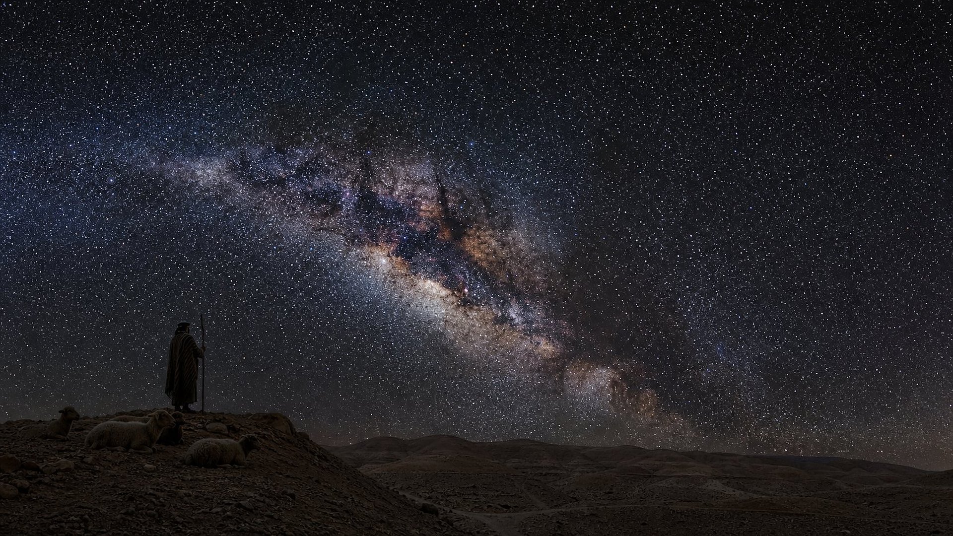 Sous un ciel nocturne sans lune, la Voie lactée forme une grande arche lumineuse de nuages d’étoiles et de bandes de poussière sombre au-dessus des collines rocailleuses du désert de Judée ; sur une crête, la silhouette d’un berger appuyé sur un bâton droit contemple le ciel, quelques moutons couchés près de lui.